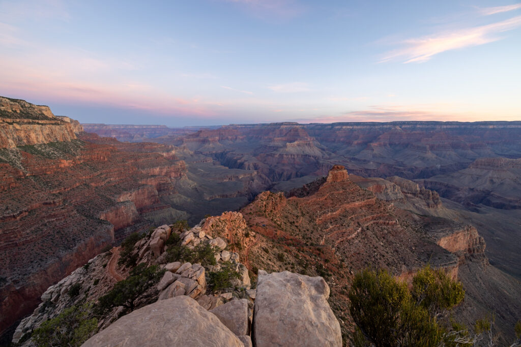 Expansive canyon landscape at sunrise from Ooh Aah Point, showing layered red rock formations, deep valleys, and distant ridges under a soft pink and blue sky