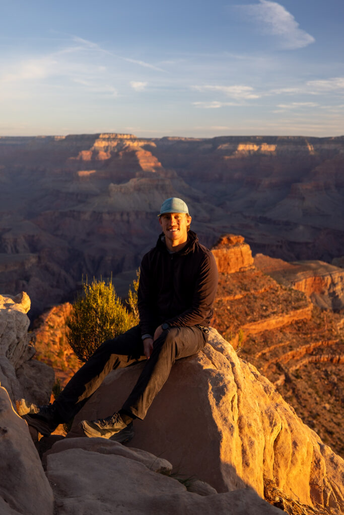 Man sitting on a rock at Ooh Aah Point during sunrise, with the Grand Canyon's layered rock formations illuminated in golden light behind him