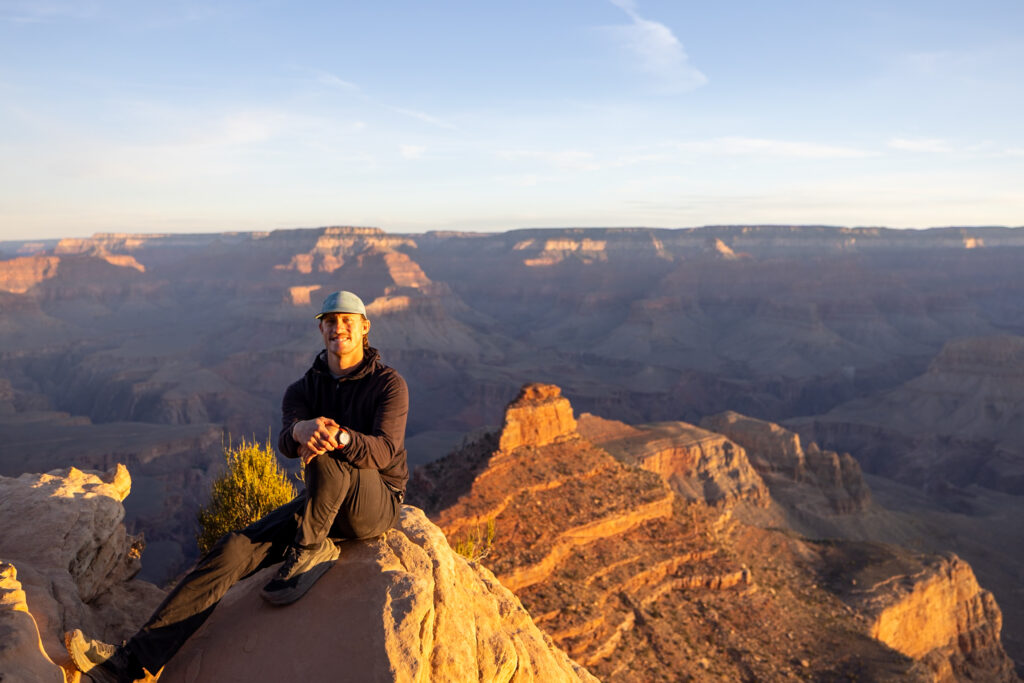 Elliot sitting on a rocky outcrop at Ooh Aah Point at sunrise, with the Grand Canyon's layered rock formations illuminated by golden morning light in the background
