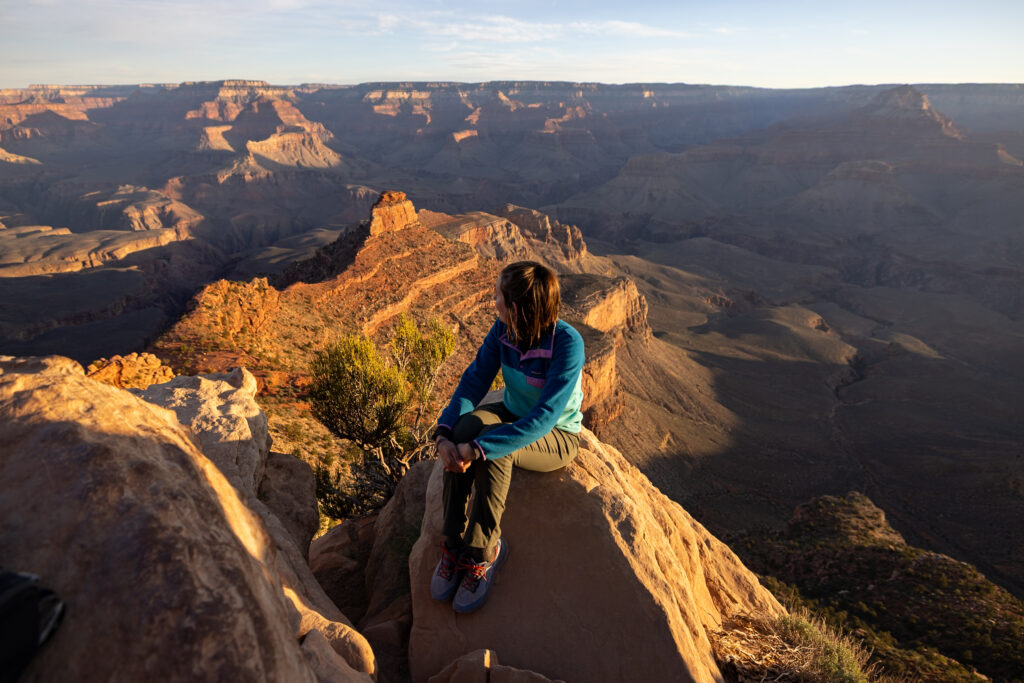 Hiker sitting on rocky outcrop at Ooh Aah Point with expansive views of the Grand Canyon's layered rock formations and distant plateaus in golden afternoon light
