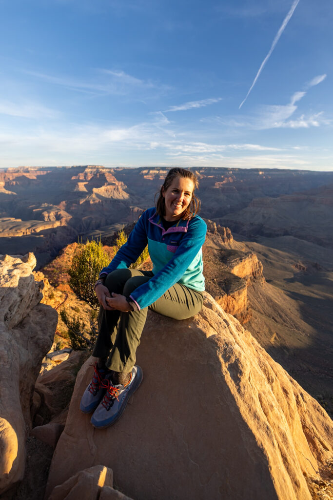 Woman sitting on a rocky outcrop at Ooh Aah Point at sunrise, with the Grand Canyon stretching behind her under a clear blue sky