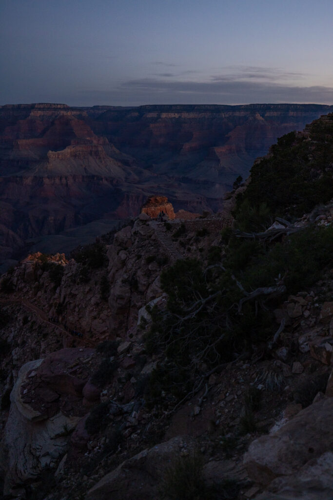 Early morning light illuminates canyon rock formations along the South Kaibab Trail, with layered canyon walls visible in the distance under a dawn sky