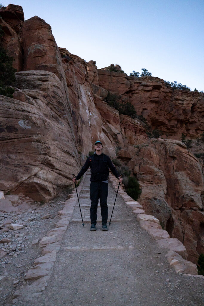 Hiker with trekking poles on the South Kaibab Trail descending toward Ooh Aah Point, surrounded by red rock canyon walls at dawn