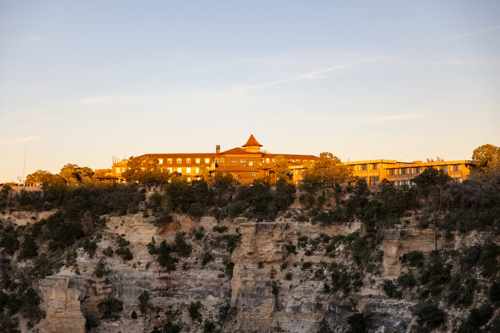 Grand Canyon Village buildings glowing in warm sunset light on the rim above the canyon's layered rock formations and vegetation