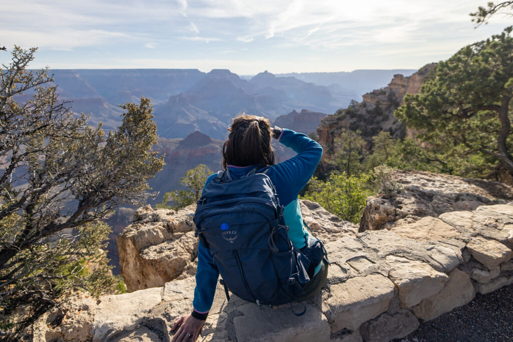 Jen in blue jacket and backpack viewing the Grand Canyon from Yaki Point, with layered canyon walls and vegetation visible in the background