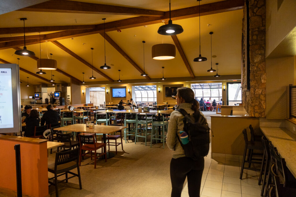 Woman standing in a spacious dining hall with warm yellow walls, wooden beams, pendant lighting, and scattered tables and chairs; other diners visible in the background