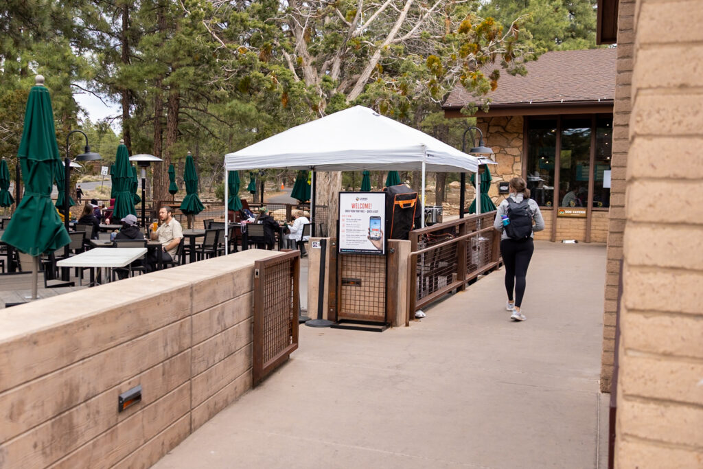 Jen walking past Yavapai Tavern's outdoor seating area with green umbrellas and dining tables surrounded by pine trees