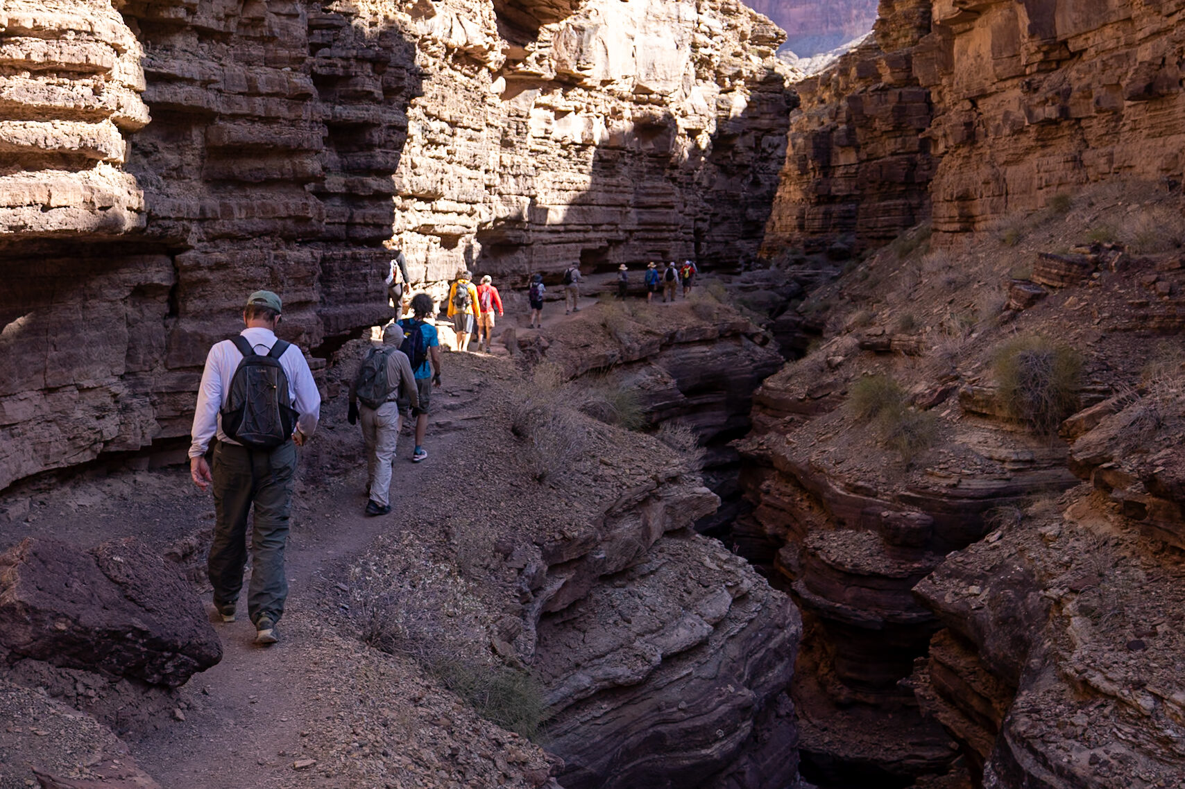 Hikers walking through a narrow canyon corridor with towering rock walls on the Deer Creek Trail, demonstrating the tight passage leading to the patio area