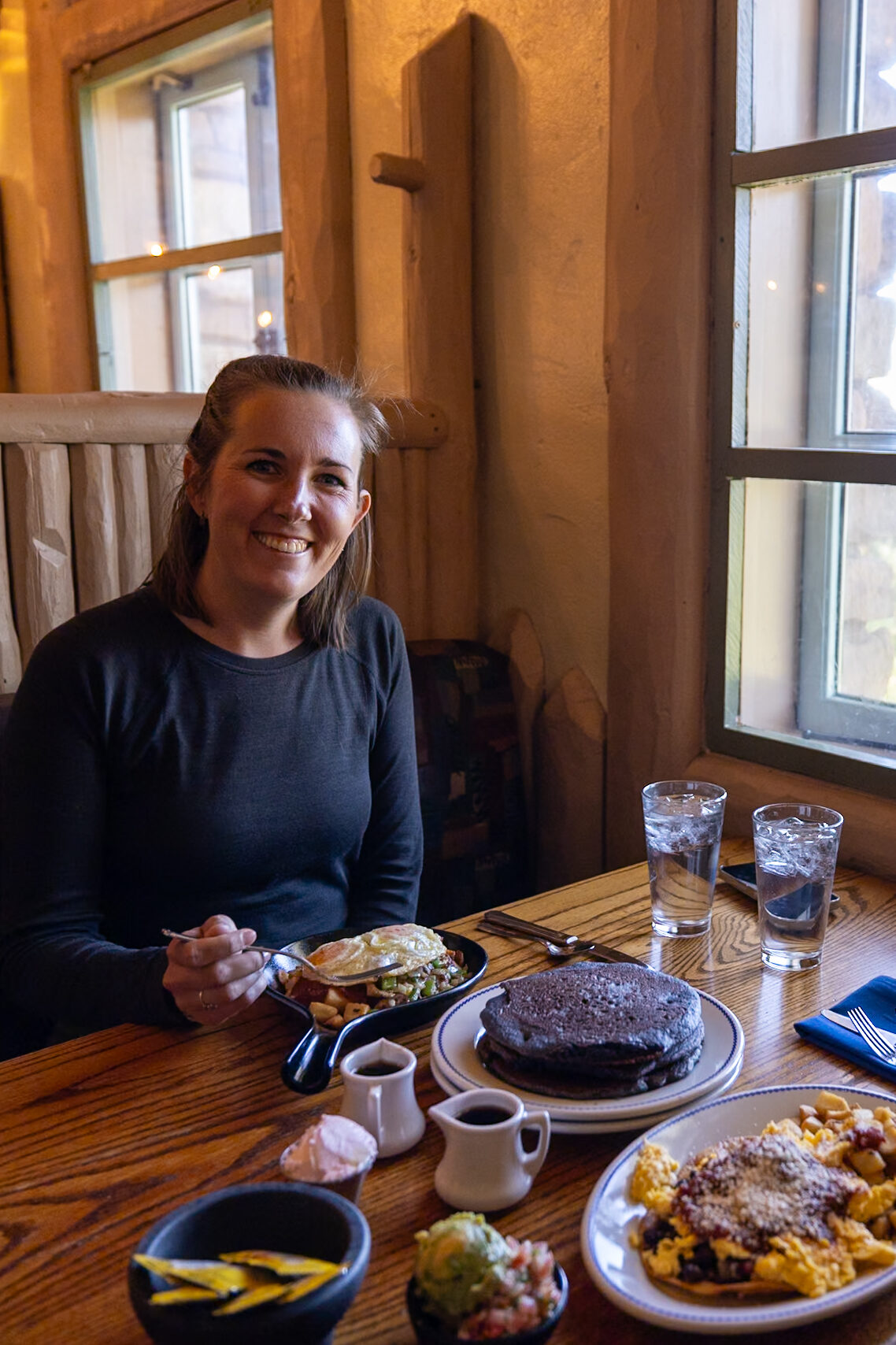 Woman dining at Harvey House Cafe with pancakes, hash browns, and eggs on the table, windows overlooking the Grand Canyon in background