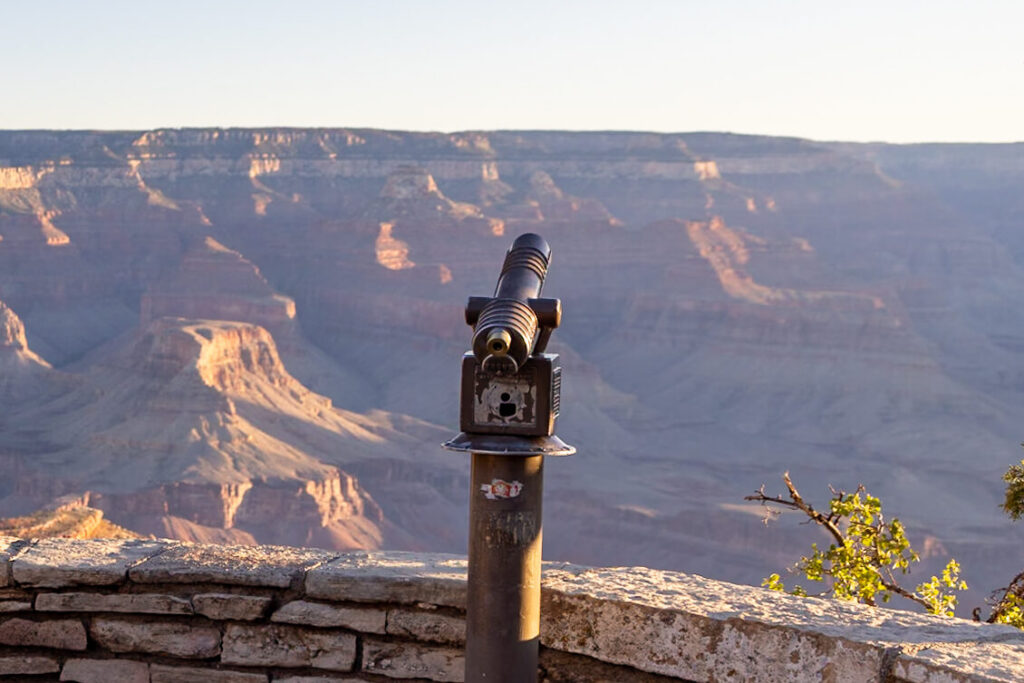 A coin-operated telescope mounted on a stone wall overlooking the Grand Canyon's layered rock formations and distant ridges. Walking the rim trail is one of the best things to do at the Grand Canyon.