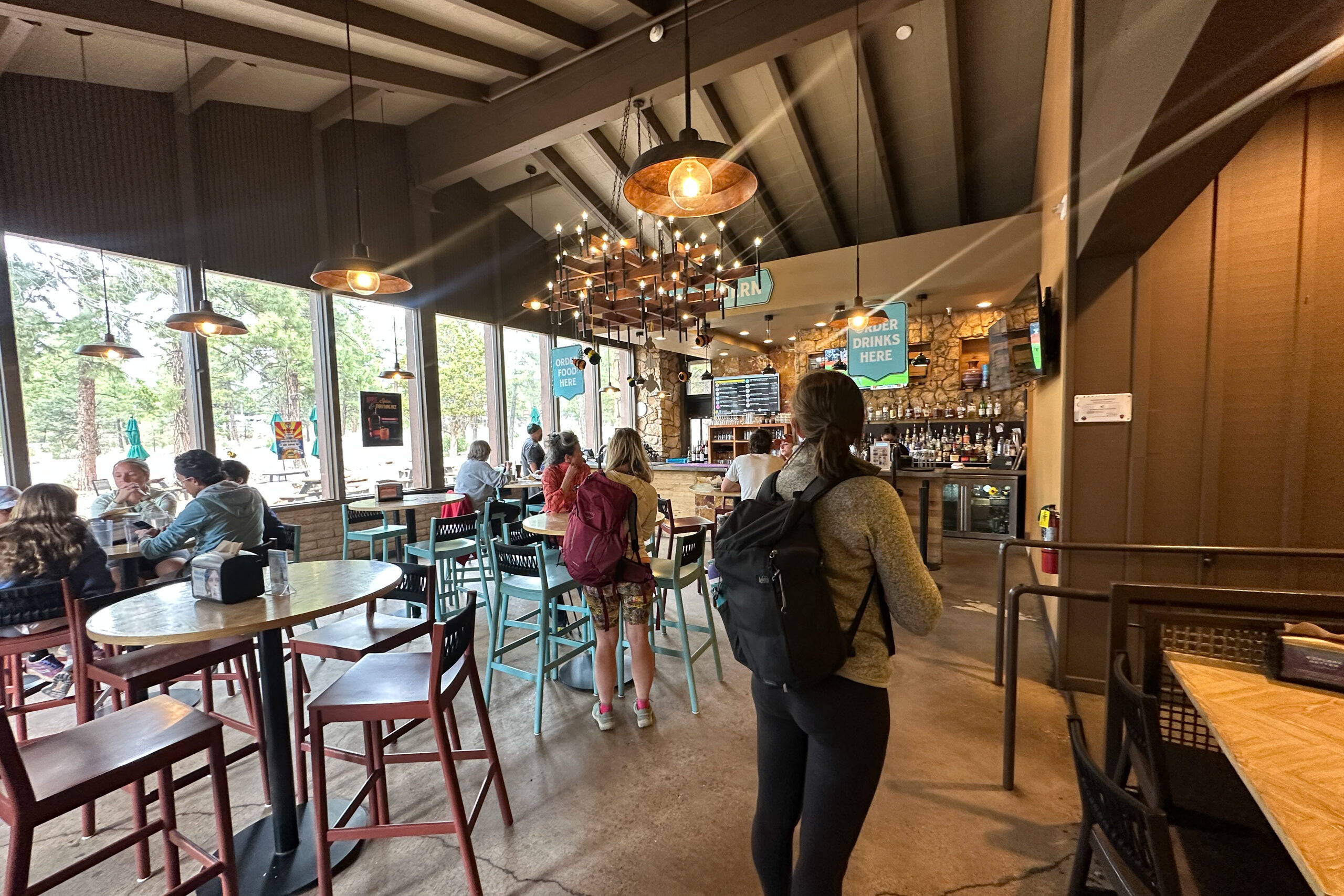 Woman standing inside Yavapai Tavern, a casual dining establishment with high ceilings, pendant lighting, a bar area, and large windows overlooking trees outside