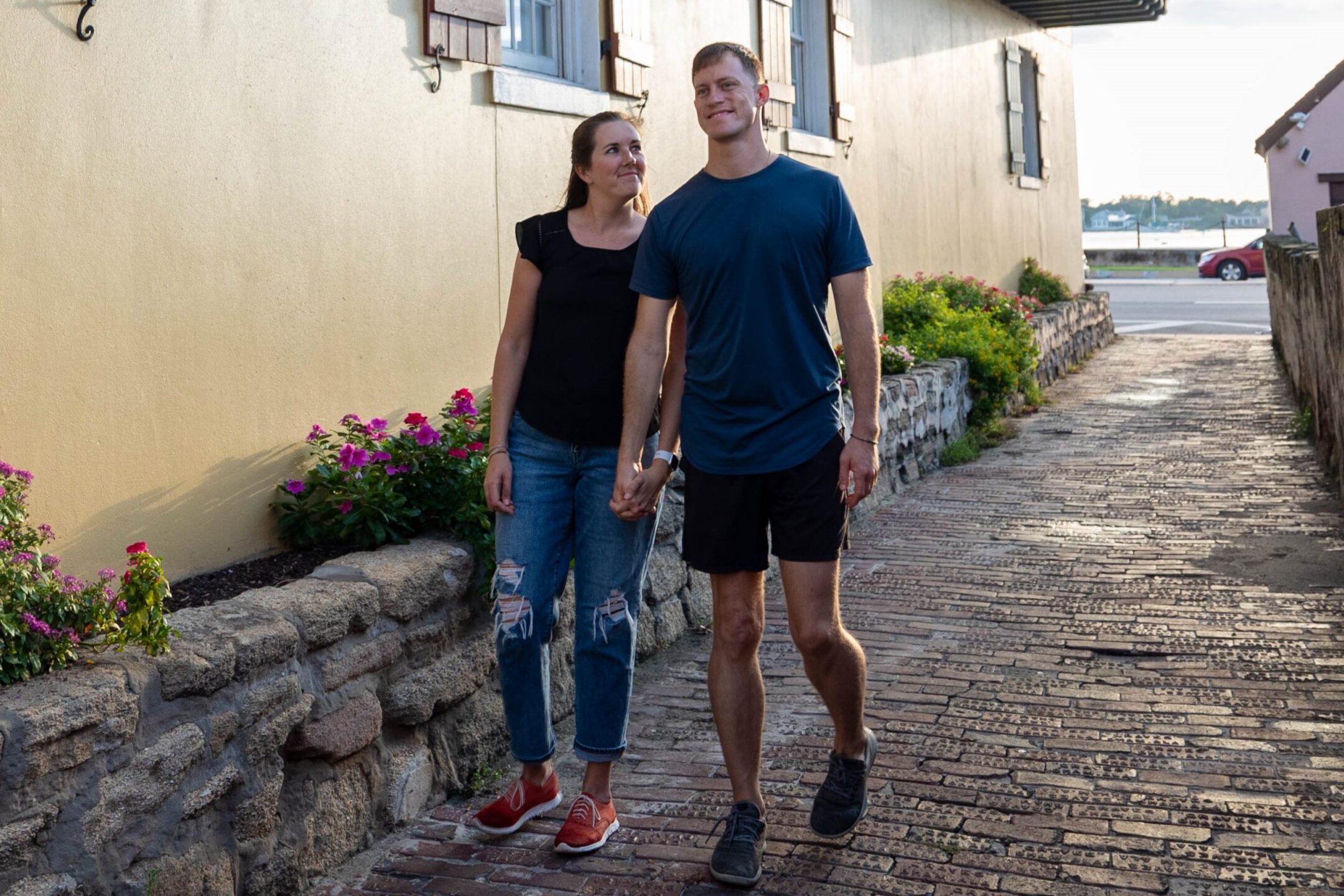 Couple walking together down a narrow brick street in historic St. Augustine, lined with cream-colored buildings and pink flowers