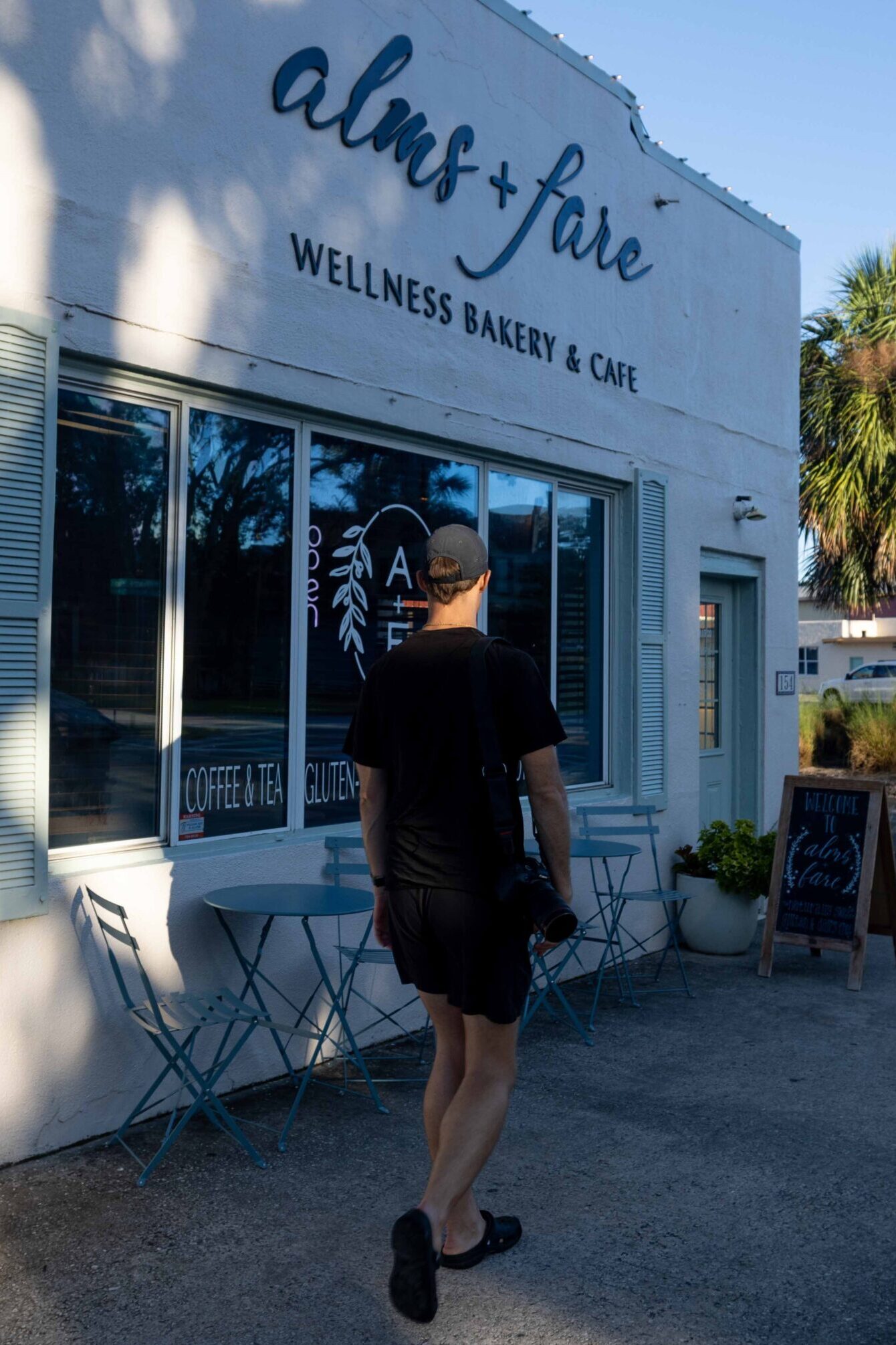 Person approaching the exterior of Alms + Fare, a white wellness bakery and cafe with large windows and outdoor seating