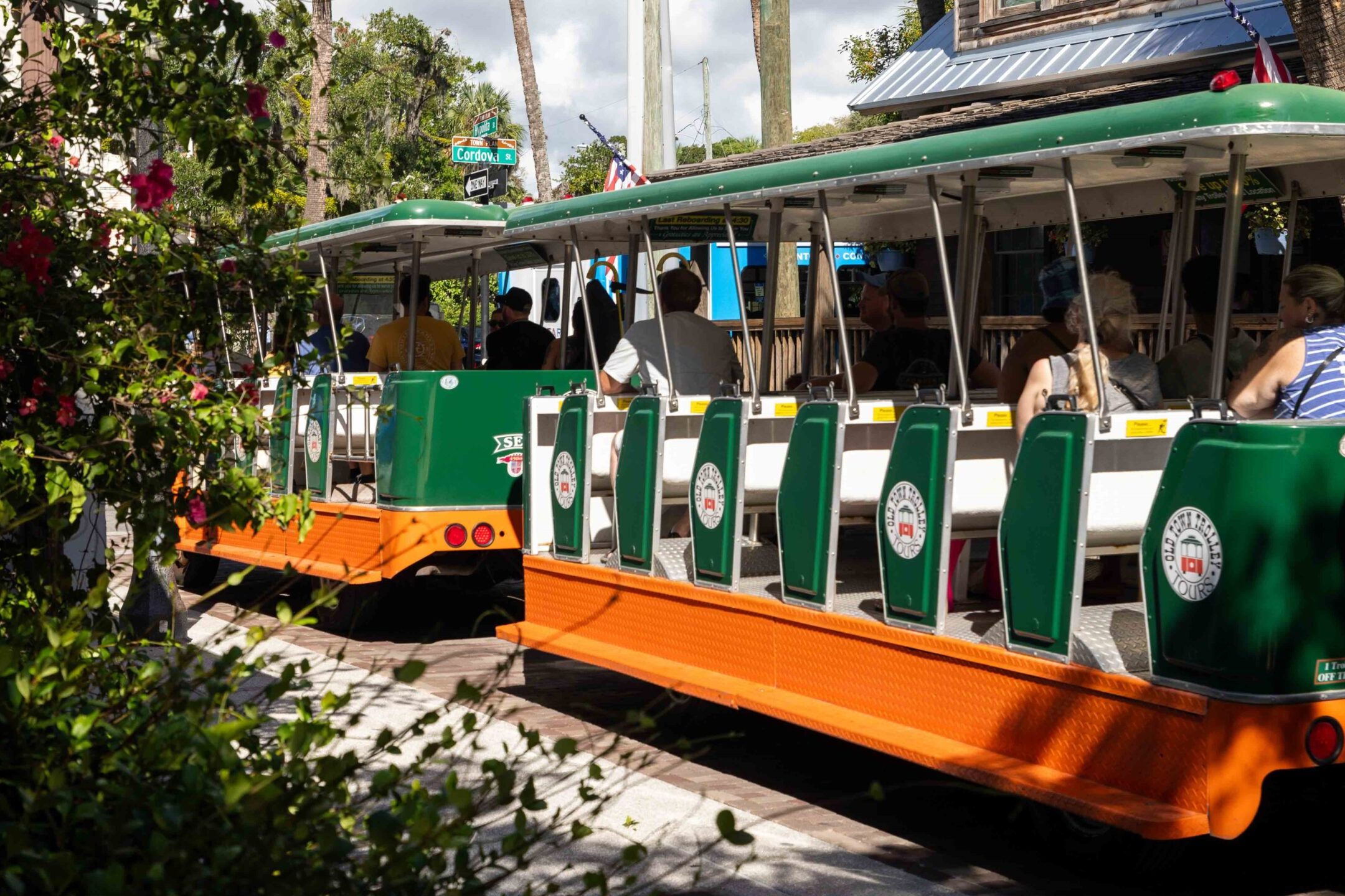 Green and orange Old Town Trolley with passengers traveling through St. Augustine's historic downtown streets lined with flowering plants and historic buildings