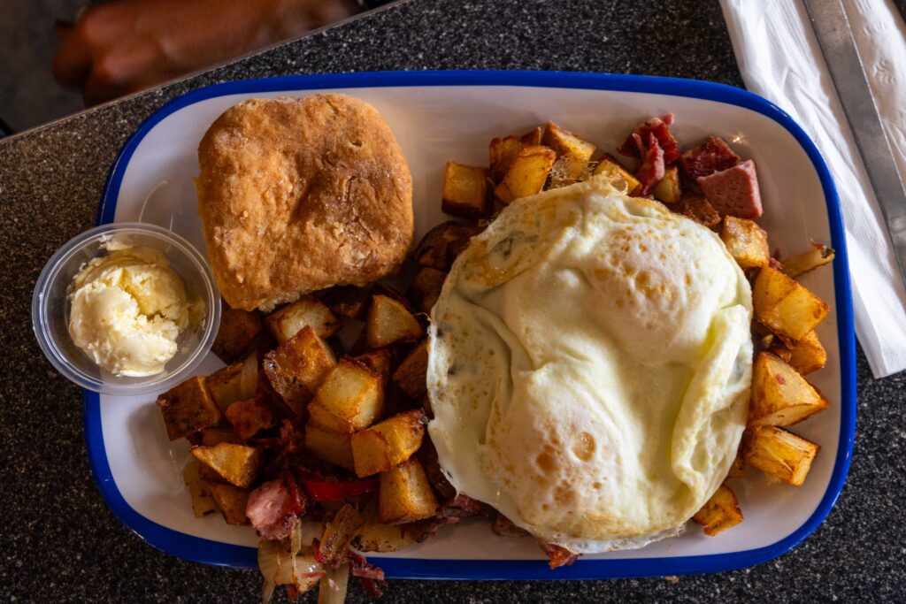 Breakfast plate with two eggs over hash browns and crispy potatoes, a biscuit, and a small container of peach butter